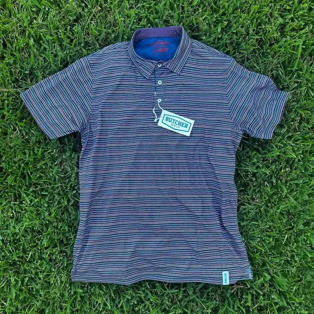 Finished custom Butcher Golf polo laid flat on grass, showing an all-over dye-sublimated repeat pattern and contrast collar.